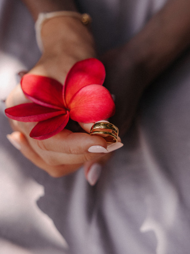 PURELEI Twin Tides Ring aus vergoldetem Edelstahl, zwei geschwungene Bänder, elegant gehalten in warmer Hand mit roter Blüte.
